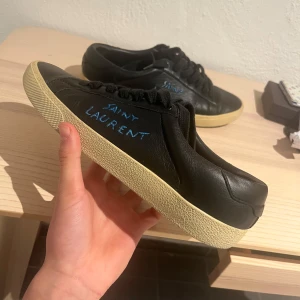 Svarta sneakers från Saint Laurent - Snygga svarta sneakers från Saint Laurent med en krämfärgad sula. Skorna har en stilren design med blå text på sidan. Perfekta för en trendig och avslappnad look. Storlek 40 men passar 41. Inget OG tillkommer. Hör av dig vid frågor eller funderingar!🤝