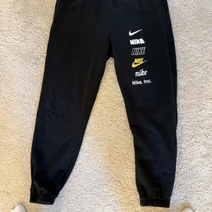 Svarta mjukisbyxor från Nike - Säljer ett par svarta mjukisbyxor från Nike med flera logotyper i vitt och gult på benet. Byxorna har en elastisk midja med snörning för justerbar passform. Storlek M. Mycket bra skick