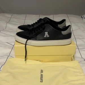 Svarta sneakers från Axel Arigato - Snygga svarta sneakers från Axel Arigato med grå detaljer och vit sula. Skorna har en stilren design med snörning och en diskret logga på sidan. Perfekta för en casual look. Knappt använda kanske använt de 5-6 ggr. Storlek 46