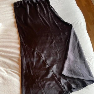 Långkjol - Elegant svart långkjok från Gina Tricot i satin. Kjolen har en enkel och stilren design, perfekt för speciella tillfällen. Den är axelbandslös och har en lätt utsvängd form som ger en vacker siluett. Den är i storlek M