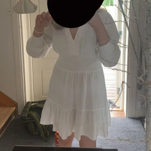 Vit klänning med volangdetaljer - Charmig vit klänning med v-ringad hals och volangdetaljer. Perfekt för sommarens festligheter. Klänningen har långa ärmar. 