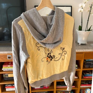 Grå hoodie med broderad rygg - Säljer en grå hoodie från Odd Molly med en unik broderad design på ryggen i svart och orange. Tröjan har en dragkedja framtill och en mysig huva. Perfekt för en avslappnad stil.