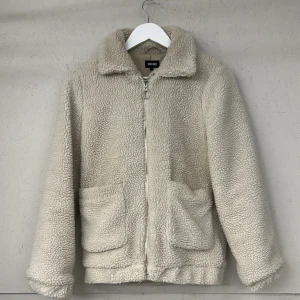 Beige teddyjacka från Bubbleroom - Mysig beige teddyjacka från Bubbleroom med dragkedja framtill och två praktiska fickor. Perfekt för kyliga dagar med sin fluffiga och varma design. Jackan har en klassisk krage och är helfodrad för extra komfort.