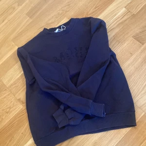 Mörkblå sweatshirt med text - Säljer en mörkblå sweatshirt med texten 'AMOUR' i pärlor på framsidan. Tröjan har långa ärmar och ribbade muddar vid ärmslut och nederkant. Perfekt för en avslappnad stil. Str 158/164