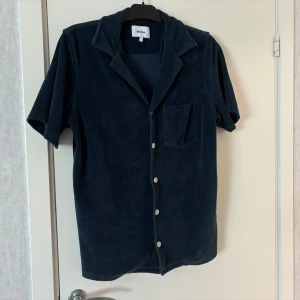 Mörkblå kortärmad skjorta/overhsirt från Nikben - Snygg mörkblå skjorta/overshirt från Nikben i frotté med korta ärmar och knappar framtill. Skjortan har en klassisk krage och en bröstficka för en avslappnad stil. Perfekt för varma dagar eller som ett stilrent lager. Den är endast använd ett fåtal gånger och skicket är 10/10 Nypris 1200
