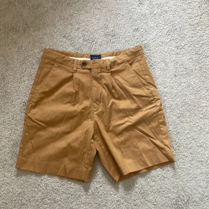 Beigea chino-shorts från Gant, storlek EU 46/ w31 - Säljer ett par stilrena beige shorts från Gant. De har en klassisk design med knappar och pressveck. Perfekta för en avslappnad och snygg look. De är ostrukna vid fototillfälle men är knappt slitna eller använda.