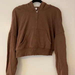 Brun croppad hoodie - En brun croppad hoodie med dragkedja och huva. Tröjan har långa ärmar och en avslappnad passform.