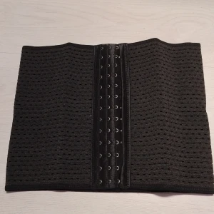Svart waisttrainer korsett med hyskor - Säljer en svart waisttrainer korsett med hyskor framtill. Den har ett perforerat mönster för bättre andningsförmåga och är tillverkad i Kina. Perfekt för att forma midjan och ge stöd. Storlek Large 74-78 cm höjd  24.5 cm