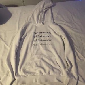 Vit hoodie från Peak Performance - Säljer en stilren vit hoodie från Peak Performance med tryck på framsidan. Tröjan har en klassisk känguruficka och en bekväm huva. Perfekt för en avslappnad stil.