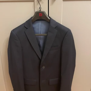 Dressman  kostym - Elegant mörkblå kostym med kavaj och matchande byxor från dressman. Kavajen har klassisk design med två knappar och fickor fram. Perfekt för formella tillfällen.