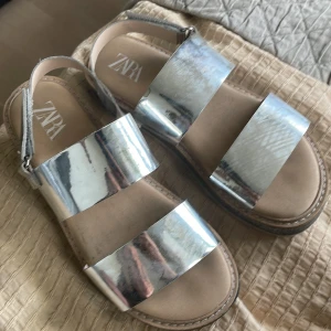 Silvriga sandaler från Zara, strl 34 - Glansiga sandaler från Zara i metallic silver med breda remmar och kardborreband runt hälen. Snygg platt sula med svart glitterdetalj på sidan. Perfekta för att sticka ut i sommarens alla looks. Det är bara att torka av dom för att de ska se helt nya ut