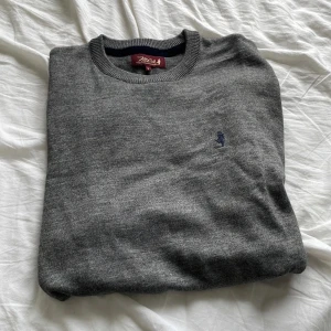 Grå meriono ull tröja - Säljer en klassisk grå stickad tröja med rund halsringning och diskret broderad logga på bröstet. Perfekt för lager-på-lager och passar till det mesta. Mjuk och bekväm med ribbade muddar vid ärmslut och nederkant.