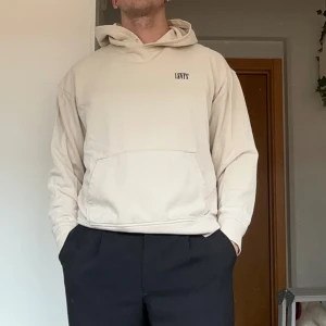 Beige hoodie från Levi's - Säljer en beige hoodie från Levi's i avslappnad passform. Tröjan har huva, magficka och diskret Levi's-logga på bröstet. Tillverkad i mjuk bomull, perfekt för chill dagar. Passar dig som gillar en clean och enkel stil.