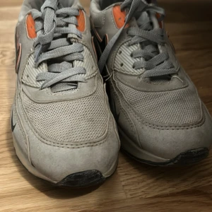 Nike Air Max 90 grå/orange sneakers - Nike Air Max 90 sneakers i grått med orange detaljer och svarta swoosh-loggor. Ovandel i mesh och syntet med klassisk Air Max-sula och snörning. Perfekt för dig som gillar retrostil och streetwear. Snygg kontrast mellan färgerna och ikonisk design.