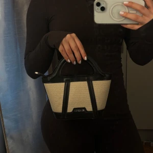 Beige och svart handväska  - Snygg handväska  i beige canvas med svarta skinn-detaljer. Väskan har en stilren, boxig form med korta handtag och logga framtill. Perfekt storlek för det viktigaste och ger en clean vibe till din outfit.