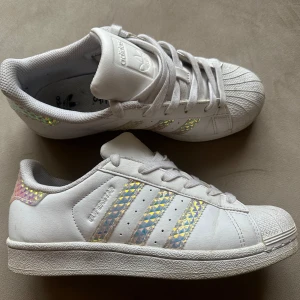 Ortholite Adidas Superstar iridescent  - Snygga vita Adidas Superstar sneakers med holografiska ränder. Klassisk snäcktå, snörning och platt sula. Perfekta för dig som vill sticka ut med en glittrig touch till din streetstyle.