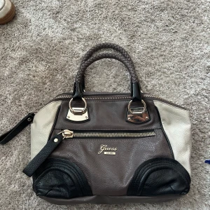 Guess handväska i skinn med dragkedja - Snygg handväska från Guess i skinn med flätade handtag. Väskan är färgblockad i grått, svart och beige med guldfärgade metalldetaljer och dragkedja framtill. Insidan har blommigt foder i blått och lila. Perfekt för dig som vill ha en stilren men unik väska.