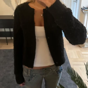 Svart stickad kofta  - Mysig svart stickad kofta utan knappar med lång ärm och rund halsringning. Koftan är enkel och stilren utan synliga knappar eller dragkedja, perfekt att slänga över en topp. Den har en lite fluffig textur som ger en cool och avslappnad vibe.