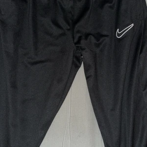 Svarta träningsbyxor från Nike - Svarta mjukisbyxor från Nike med klassisk vit swoosh-logga på låret. Byxorna har en sportig look och är tillverkade i ett glansigt syntetmaterial som känns lätt och smidigt. Perfekta för träning eller chill. Resår i midjan för skön passform. Skador på vänstra sidan vid vadbenet och det är riven skada och hål . Ingen Frakt
