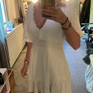 Vit klänning  - Supersöt vit kortklänning från Amisu med v-ringning och spetsdetalj vid urringningen. Klänningen har korta, vida ärmar och markerad midja med smock. Volangig kjol ger en flowy känsla, perfekt för sommarens alla tillfällen.