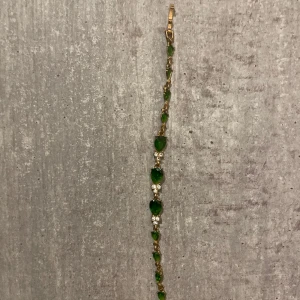 Guldarmband med gröna stenar - Ett elegant armband i guldton med droppformade gröna stenar och små gnistrande vita stenar mellan. Armbandet har en klassisk låsning och ger en lyxig känsla med sina färgstarka detaljer. Perfekt för dig som vill sticka ut med stilrena smycken.