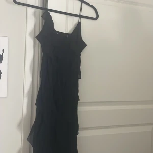 Svart långklänning med volang - Svart långklänning med tunna axelband och volangdetaljer längs sidorna. Klänningen har en luftig och flowy känsla, perfekt för dig som gillar en elegant och stilren look. Materialet är lätt och ger ett snyggt fall.