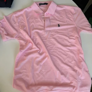 Rosa pikétröja från Ralph Lauren - Säljer en klassisk ljusrosa pikétröja från Ralph Lauren med broderad logga på bröstet. Tröjan har korta ärmar, krage och knäppning med två knappar. Perfekt för en fräsch och sportig look.
