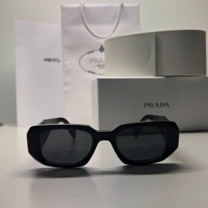 Svarta solglasögon från Prada - Populära solglasögon från Prada med rektangulära, kantiga bågar och mörka glas. Tjocka skalmar med Prada-logga på sidan. Knappt använda, väldigt bra skick. Allt original förpackning samt kvitto medföljer. 