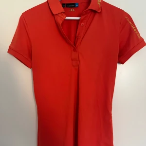 Röd pikétröja från J.Lindeberg - Snygg röd pikétröja från J.Lindeberg med klassisk krage och knappar framtill. Tröjan har korta ärmar och gul logga på ärmen och nacken. Tillverkad i mjuk polyester som andas, perfekt för en sportig och stilren look.