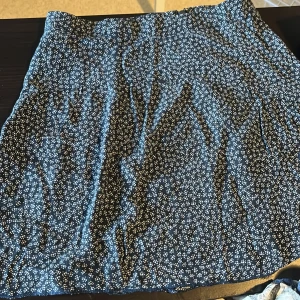 Mörkblå blommig kortkjol - Supersöt svart kortkjol med små vita blommor över hela tyget. Kjolen har en enkel och rak passform som är lätt att matcha med olika toppar. Perfekt för dig som gillar en chill och trendig stil.