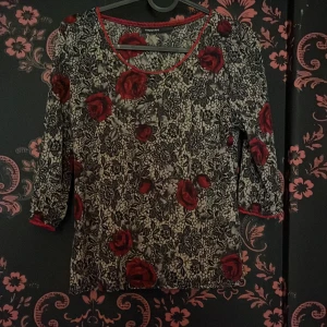 Blommig topp från KappAhl med röda rosor - Supersnygg topp från KappAhl med svartvitt spetsinspirerat mönster och stora röda rosor. Rund halsringning med röd spetskant och trekvartsärm. Perfekt för dig som gillar romantiska vibes och vill sticka ut lite extra.