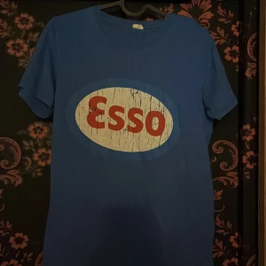 Blå Esso t-shirt med tryck - Snygg blå t-shirt med klassiskt Esso-tryck i rött och vitt framtill. T-shirten har rund hals och korta ärmar, perfekt för en avslappnad och cool stil. Passar dig som gillar retro och streetwear.