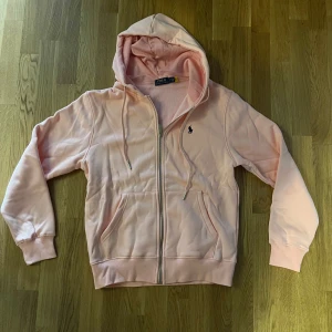 Ljusrosa hoodie från Polo Ralph Lauren - Säljer en ljusrosa hoodie från Polo Ralph Lauren med dragkedja och huva. Tröjan har långa ärmar, snörning i huvan och klassisk blå logga broderad på bröstet. Mjuk insida och två stora fickor framtill. Perfekt för chill dagar. Oanvänd, pris kan diskuteras. L men sitter som M