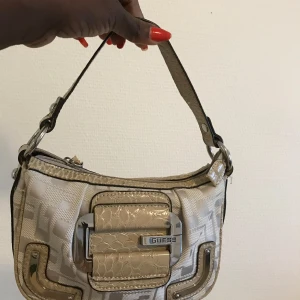 Beige axelväska från Guess - Snygg beige axelväska från Guess med glansiga detaljer och croco-mönstrad rem. Väskan har ett stort metallspänne framtill med Guess-logga och dragkedja upptill. Insidan är fodrad med mönstrat tyg och har en liten innerficka med logga.