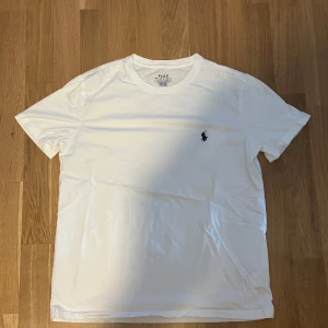 Vit t-shirt från Polo Ralph Lauren - Vit t-shirt från Polo Ralph Lauren med klassisk rund hals och broderad marinblå logga på bröstet. Slim fit-modell i mjuk bomull som sitter snyggt och passar till allt. Enkel och stilren design som alltid funkar.