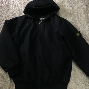 Svart vindjacka från Stone Island - Svart vindjacka från Stone Island med huva och klassisk logga på ärmen. Jackan har dragkedja framtill, ribbade muddar och två fickor med knappar. Insidan är quiltad för extra komfort. Perfekt för dig som gillar streetwear och snygg design.