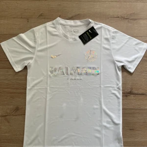 Vit PSG x Balmain fotbollströja Nike S - Snygg vit fotbollströja från Nike i samarbete med PSG och Balmain. Tröjan har korta ärmar, rund hals och är gjord i lätt, andningsbart material. Framsidan pryds av holografiska loggor och text. Perfekt för dig som vill sticka ut på planen eller gatan.