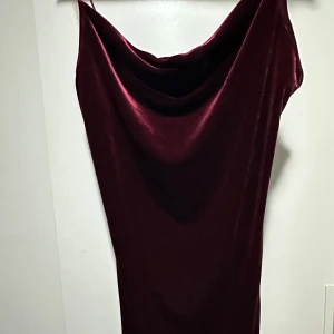 Vinröd sammetsklänning med draperad urringning - Säljer en vinröd långklänning i mjuk sammet med draperad urringning och smala axelband. Klänningen har en elegant, figurnära passform och är perfekt för dig som vill sticka ut med en lyxig känsla. Stilren och tidlös design som ger en snygg siluett.