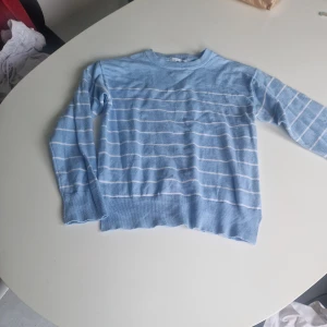 Ljusblå randig tröja från 157 - Säljer en ljusblå stickad tröja från 157 med vita horisontella ränder. Tröjan har rund halsringning, långa ärmar och ribbade muddar vid hals och nederkant. Perfekt för dig som gillar en chill och stilren look.