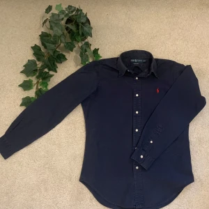 Mörkblå skjorta från Ralph Lauren  - Klassisk mörkblå skjorta från Ralph Lauren i bomull med lång ärm och vit knappstängning. Broderad röd logga på bröstet och klassisk krage. Modellen är Classic Fit och passar perfekt till en stilren look.