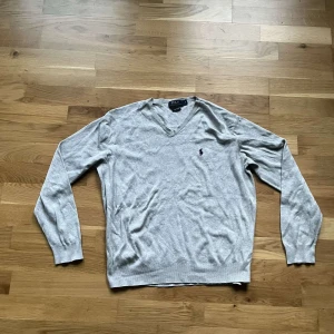 Ralph Lauren tröja  - Snygg ljusgrå v-ringad tröja från Polo Ralph Lauren i mjuk bomull. Klassisk passform med långa ärmar och broderad logga på bröstet. Perfekt att slänga på sig över en t-shirt eller skjorta för en clean look.