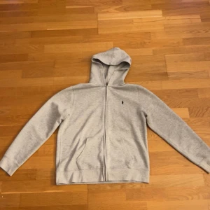 Grå hoodie från Ralph Lauren - Snygg ljusgrå hoodie från Ralph Lauren med dragkedja framtill och klassisk huva. Tröjan har två fickor och den ikoniska lilla loggan broderad på bröstet. Perfekt för chill dagar eller när du vill ha en enkel och stilren look.