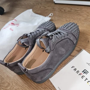 Gråa Louboutins - Snygga grå sneakers i mocka från Christian Louboutin med coola nitar över tån (2 nitar saknas).Skorna har snörning och en rund tå, samt en platt sula. Perfekta för dig som vill sticka ut med en edgy och lyxig stil. 