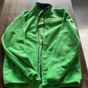 Grön fleecejacka från Clique L - Snygg grön fleecejacka från Clique i storlek L. Jackan har hel dragkedja framtill, hög krage och två sidofickor med dragkedja. Perfekt för kyliga dagar när du vill vara både bekväm och stilren.