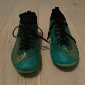 Nike Mercurial turkosa fotbollsskor CR7 - Säljer ett par Nike Mercurial fotbollsskor i turkos med svarta och guldiga detaljer. Skorna har snörning, högre strumpliknande skaft och orange dobbar för gräsplan. Snygg design med guldigt mönster på sidan och Mercurial-tryck bak och ny tvättade men har en liten spricka under. Har varit använda en halv fotbolls säsong och blev lite för små för mig. Frakt 50kr