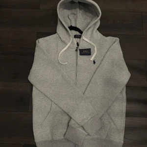 Grå hoodie från Polo Ralph Lauren - Snygg ljusgrå hoodie från Polo Ralph Lauren med dragkedja och vit snörning i huvan. Klassisk broderad logga på bröstet och mjukt material som känns skönt mot huden. Perfekt för chill dagar eller när du vill ha en clean streetstyle.