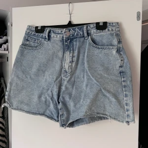 Ljusblåa jeansshorts - Ljusblåa jeansshorts med hög midja och avslappnad passform. Klassisk femficksmodell med råa, fransiga benslut. Perfekta för varma dagar och enkel att matcha med allt. Dragkedja och knapp framtill, bälteshällor och bakfickor. Önskar du veta exakta mått? Tveka inte att höra av dig!