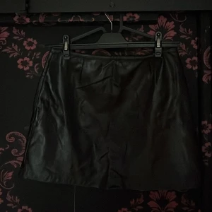 Svart skinnkjol från Hennes Collection - Snygg svart kort skinnkjol från Hennes Collection. Klassisk rak modell med slät yta och stilren look. Perfekt för dig som vill ha en edgy vibe till outfiten. Kjolen är enkel att matcha och har en tidlös känsla.