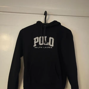 Svart hoodie från Polo Ralph Lauren - Svart hoodie från Polo Ralph Lauren med vit broderad logga på bröstet. Klassisk modell med huva och snörning samt känguruficka framtill. Perfekt för en chill och stilren look.