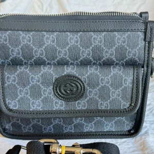 Gucci svart axelväska med GG-mönster - Snygg svart axelväska från Gucci med klassiskt GG-mönster i grått. Väskan har justerbar axelrem, dragkedja upptill och praktiskt ytterfack med lock. Tillverkad i canvas och skinn med silverfärgade metalldetaljer. Perfekt för dig som gillar stilrena designer och logomönster.